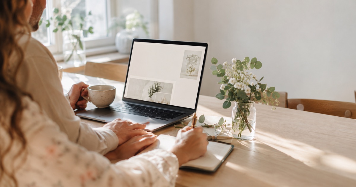 Par som planerar sitt bröllop vid ett soligt matbord med öppen laptop, anteckningsbok och eucalyptus i en vas